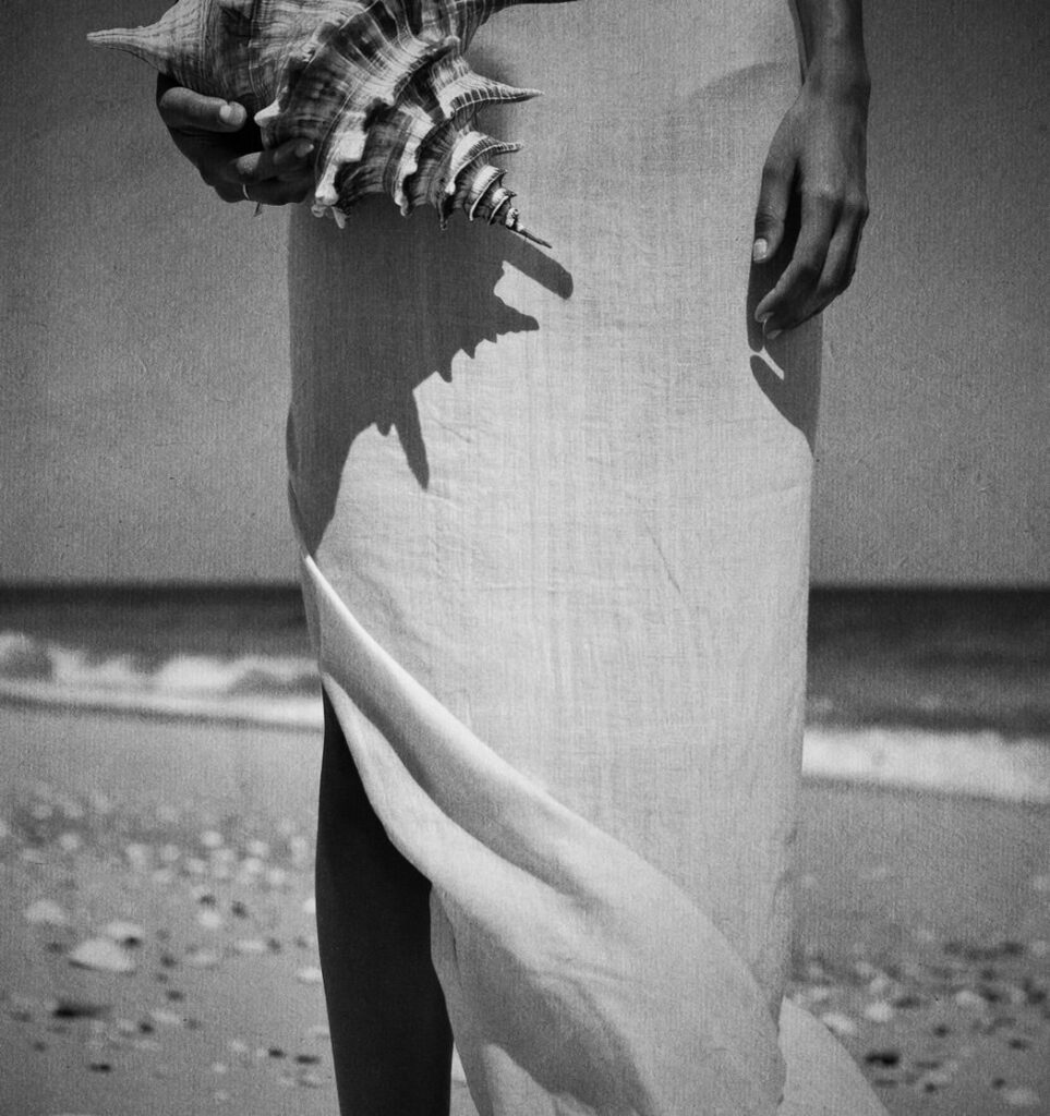A woman standing on the beach holding a seashell in her hand, with soft light and ocean tones in the background.